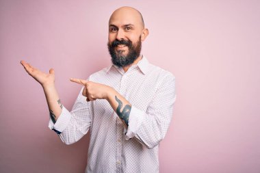 Soyutlanmış pembe arka plan üzerinde zarif bir gömlek giyen sakallı yakışıklı kel adam kameraya hayret ediyor ve gülümsüyor..