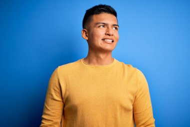 İzole edilmiş mavi arka plan üzerinde sarı günlük süveter giyen genç, yakışıklı latin adam yüzünde gülümsemeyle, doğal bir ifadeyle yan yana bakıyor. Kendinden emin gülen.