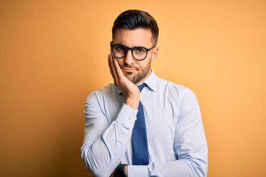 Sarı arka plan üzerinde duran kravat ve gözlük takan genç yakışıklı işadamı kollarını kavuşturarak depresyondan sıkılmış ve yorgun görünüyor..