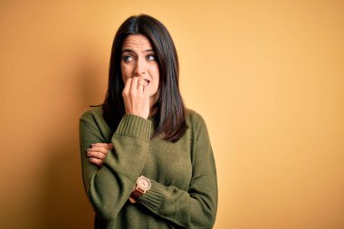 Sarı arka planda yeşil süveter giyen, mavi gözlü genç esmer kadın elleri ağızdan tırnak yerken gergin ve stresli görünüyor. Endişe sorunu.