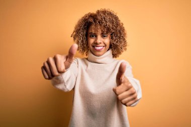 Sarı arka plan üzerine balıkçı yaka kazak giyen genç ve güzel Afro-Amerikan kadın elleriyle olumlu jest yapıyor, gülümsüyor ve başarı için seviniyor. Kazanan jest.