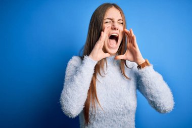 Genç, güzel, kızıl saçlı bir kadın izole edilmiş mavi arka plan üzerine günlük süveter giyip elleri ağzında kızgın bir şekilde bağırıyor.