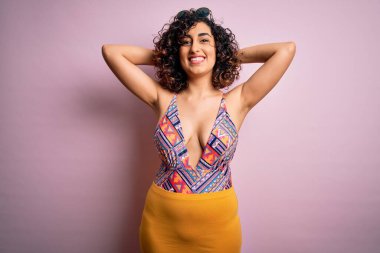 Genç, güzel bir Arap kadın, mayo ve güneş gözlüğü takmış pembe arka planda dinleniyor ve esniyor, kolları ve elleri başının arkasında mutlu bir şekilde gülümsüyor.