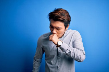 İzole edilmiş mavi arka plan üzerinde dikilen ve soğuk algınlığı veya bronşitin belirtisi olarak öksüren genç, yakışıklı Çinli bir adam. Sağlık hizmeti kavramı.