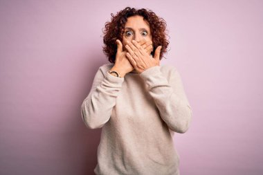 Orta yaşlı, kıvırcık saçlı, pembe arka planda balıkçı yaka kazak giyen güzel bir kadın ağzını yanlışlıkla elleriyle kapatarak şok etti. Gizli konsept.