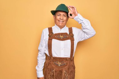 Sarı arka planda Alman geleneksel ahşap şenliği kıyafeti giyen yaşlı gri saçlı adam el ve parmaklarıyla tamam işareti yaparken gülümsüyor. Başarılı ifade.