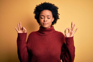 Kıvırcık saçlı, balıkçı yaka kazak giyen, gözleri kapalı gülümseyen ve meditasyon yaparken parmaklarıyla gülümseyen güzel Afro-Amerikan kadın. Yoga konsepti.