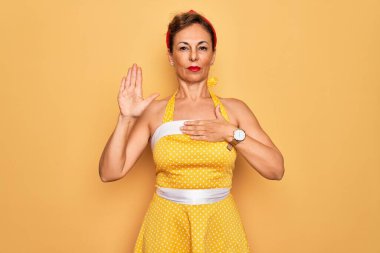 Orta yaşlı bir kadın, 50 'li yaşlarda, sarı arka planda eski tarz bir elbise giyiyor. Göğüs ve avuç içleri açık yemin ediyor, sadakat yemini ediyor.