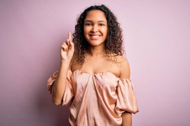 Kıvırcık saçlı, rahat tişörtlü pembe arka planda duran genç ve güzel bir kadın bir numarayı gösterirken kendinden emin ve mutlu bir şekilde gülümsüyor..