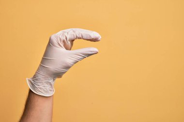 Sarı arka plan üzerinde tıbbi eldiveni olan beyaz bir gencin eli. Görünmez bir şeyi alıp götürüyor. Parmakları yer gösteren bir nesneyi tutuyor.