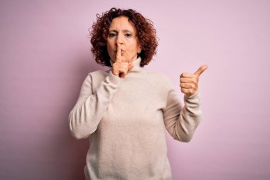 Orta yaşlı, kıvırcık saçlı, pembe arka plan üzerine balıkçı yaka kazak giyen güzel bir kadın parmaklarını yan tarafı işaret eden dudaklarıyla sessiz olmak istiyor. Sessizlik ve gizli konsept.