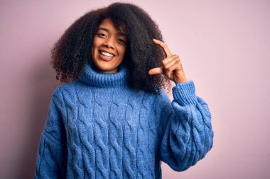 Afro saçlı, pembe arka planda kışlık süveter giyen genç ve güzel Afro-Amerikan kadın gülümseyen ve kendine güvenen, el hareketleriyle küçük boy işareti yapan, parmakları ve kamerayla bakan. Ölçü kavramı.