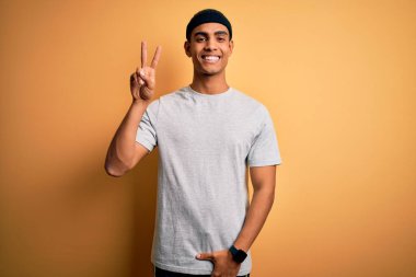 Yakışıklı Afro-Amerikan sporcusu sarı arka planda spor giysisi giyiyor gülümsüyor parmaklarını zafer işareti yaparken gösteren kameraya bakıyor. İki numara..