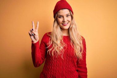 Genç, sarışın, sıradan bir kazak giyen, beyaz arka planda yün şapkalı, gülümseyen, parmaklarını zafer işareti yaparken gösteren kameraya bakan güzel bir kadın. İki numara..