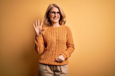 Orta yaşlı güzel sarışın kadın sarı arka planda günlük süveter ve gözlük takıyor ve parmaklarıyla beş numarayı gösterirken kendinden emin ve mutlu bir şekilde gülümsüyor..