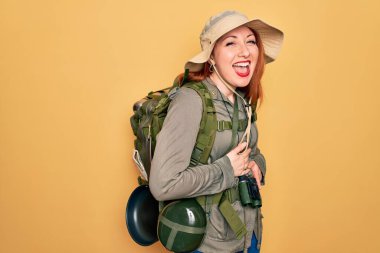 Sırt çantalı, sarı arka planda şapka takmış, sırtüstü sırtüstü gülen ve kahkahalarla gülen genç, kızıl saçlı bir kadın..