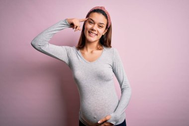 Genç, güzel bir kız hamile, pembe arka planda bebek bekliyor. Tek parmağıyla gülümseyerek, harika bir fikir ya da düşünceyle, iyi bir hafızayla.