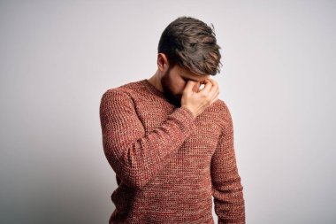 Sakallı, mavi gözlü, beyaz arka planda sıradan bir süveter giyen, burnu ve gözleri ovuşturan yorgun, bitkin ve baş ağrısı çeken genç sarışın bir adam. Stres ve hüsran kavramı.