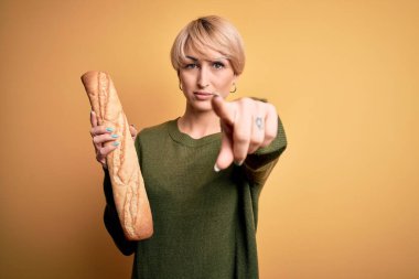 Sarı zemin üzerinde taze ekmek ekmeği tutan genç sarışın kadın kamerayı işaret ediyor ve sen, el işareti, ön taraftan pozitif ve kendinden emin bir jest.