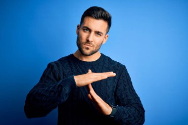 Mavi arka plan üzerinde duran sıradan bir süveter giyen genç yakışıklı adam elleri ve ciddi yüzüyle jest yapıyor.