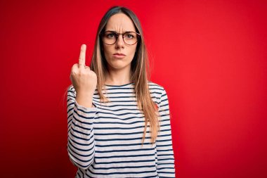 Young beautiful blonde woman with blue eyes wearing glasses standing over red background Showing middle finger, impolite and rude fuck off expression