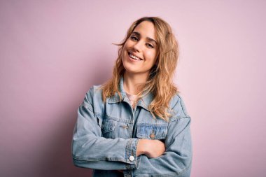 Genç, güzel, sarışın bir kadın pembe arka planda dikilmiş kameraya bakarken kollarını çapraz yapıp gülümseyen sıradan bir kot ceket giyiyor. Pozitif kişi.