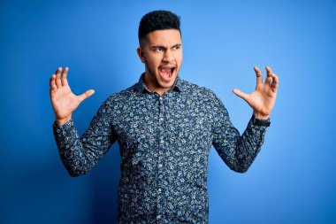 Genç ve yakışıklı bir adam yalnız mavi arka planda dikiliyor çılgın ve kızgın bir şekilde bağırıyor ve agresif bir ifade ve kollarla bağırıyor. Hayal kırıklığı konsepti.