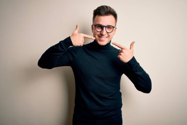 Gözlük takan yakışıklı beyaz bir adam izole edilmiş arka planda rahat bir süveter giyiyor neşeli gülümsüyor ve parmaklarını ve ağzını işaret ediyor. Diş sağlığı konsepti.