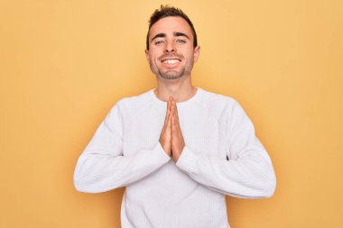 Mavi gözlü genç adam sarı arka planda dikilmiş ellerini birleştirip af diliyor kendinden emin bir şekilde gülümsüyor..