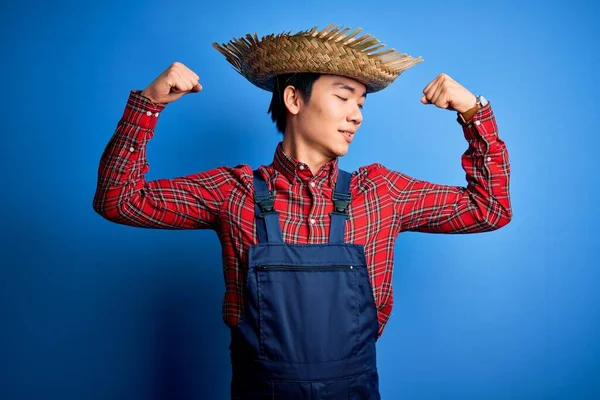 Young handsome chinese farmer man wearing apron and straw hat over blue background showing arms muscles smiling proud. Fitness concept.