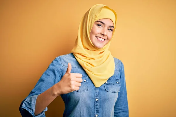 年轻美丽的女孩戴着穆斯林头巾 站在孤立的黄色背景上 手举着快乐的大拇指 在镜头前看着成功的表情 — 图库照片