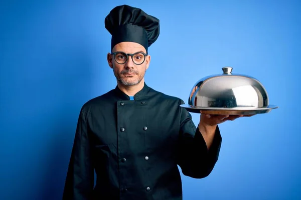 年轻英俊的厨师 身穿制服 头戴礼帽 拿着圆顶餐盘 脸上带着自信的表情 一副严肃严肃的样子 — 图库照片