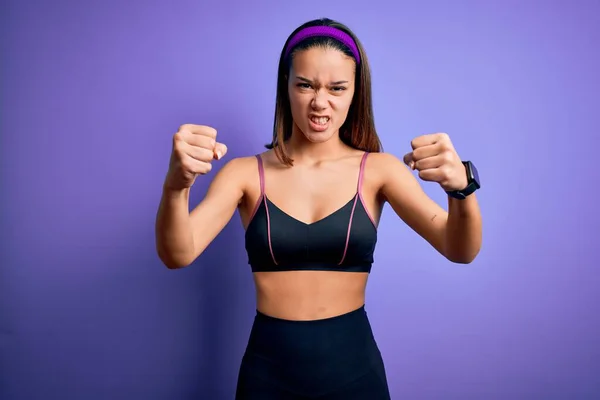年轻漂亮的女运动员穿着运动服 穿着孤立的紫色背景的运动服 怒气冲冲地举起拳头 怒气冲冲地大叫着 愤怒和好斗的概念 — 图库照片