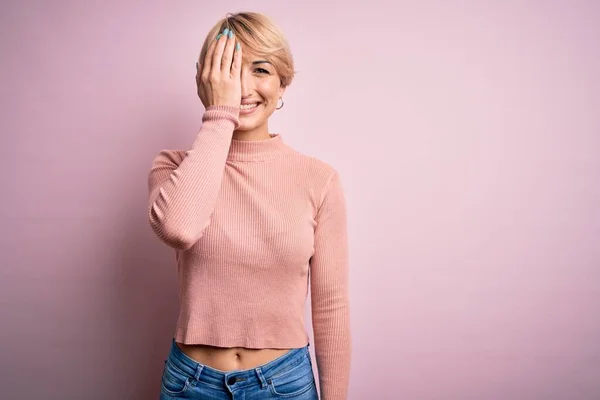 若いですブロンド女性とともに短いです髪身に着けていますカジュアルTurtleeckセーター上のピンクの背景カバー1目に手で 自信のある笑顔と驚きの感情 — ストック写真