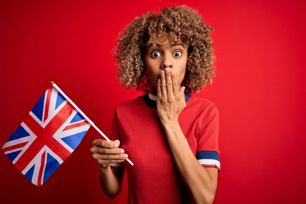 Young african american curly woman holding uk flag celebrating brexit referendum cover mouth with hand shocked with shame for mistake, expression of fear, scared in silence, secret concept