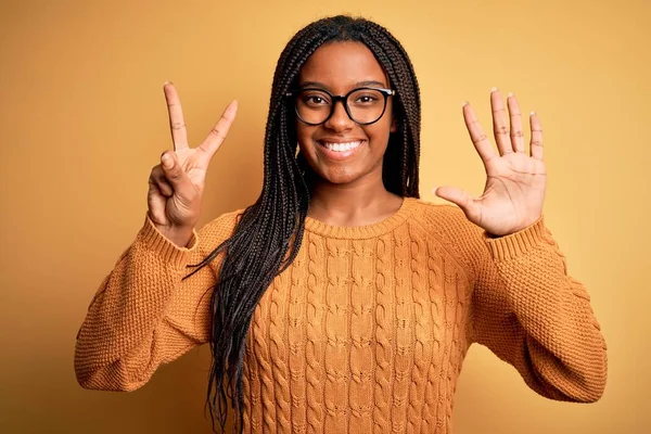 若いですアフリカ系アメリカ人スマートな女性身に着けています眼鏡とカジュアルセーター上の黄色の背景表示と指摘指番号7ながら笑顔自信と幸せ — ストック写真
