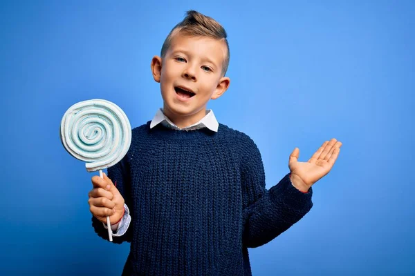 青の孤立した背景に甘いキャンディーロリポップを食べる若い小さな白人の子供は非常に幸せと興奮し 勝者の式は大きな笑顔で叫んで勝利を祝うと手を上げ — ストック写真