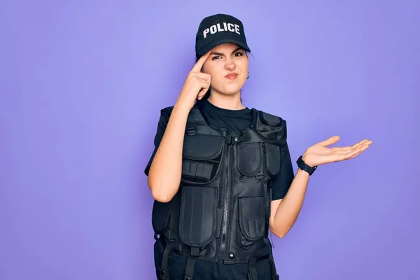 Young police woman wearing security bulletproof vest uniform over purple background confused and annoyed with open palm showing copy space and pointing finger to forehead. Think about it.