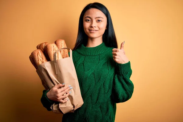 年轻美丽的中国女人拿着装有面包的纸袋 披着孤立的黄色底色 开心地笑着做着好的手势 用手指竖起大拇指 很好的标志 — 图库照片