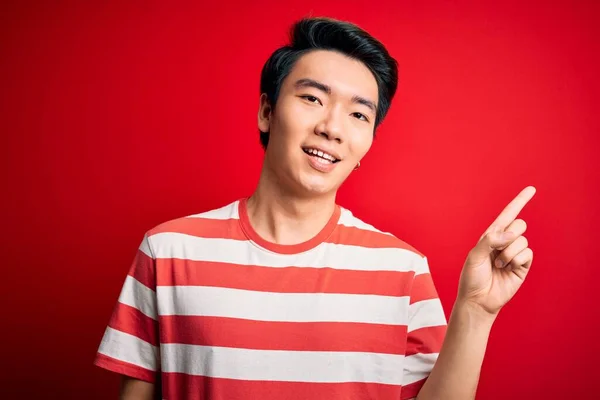 若いハンサムな中国人男性が成功した考えと指を指す赤い背景の上に立ってカジュアルな縞のTシャツを着ている 興奮し 第一番 — ストック写真