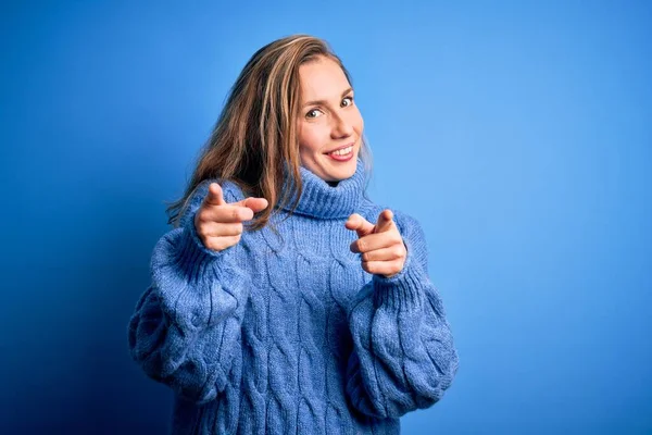 若い美しいブロンドの女性は 幸せと面白い顔を持つカメラに指を指している青い背景にカジュアルなタートルネックセーターを着ています 良いエネルギーと振動 — ストック写真