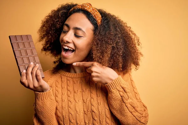 Young african american woman with afro hair holding chocolate bar over yellow background very happy pointing with hand and finger