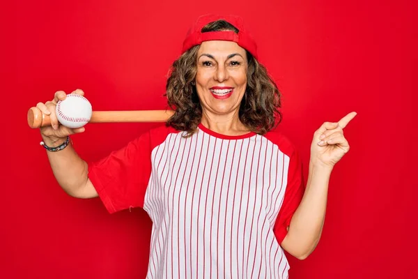 Middle age senior woman wearing baseball equiment, bat and ball over red isolated background very happy pointing with hand and finger to the side