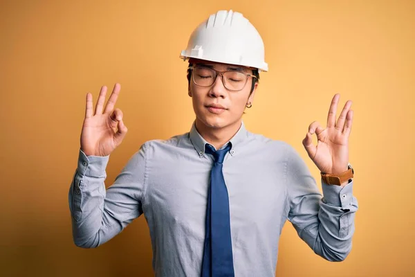 若いハンサムな中国の建築家の男は 安全ヘルメットを着用し 黄色の背景にネクタイをリラックスし 指で瞑想のジェスチャーを行う目を閉じて笑顔 ヨガのコンセプト — ストック写真