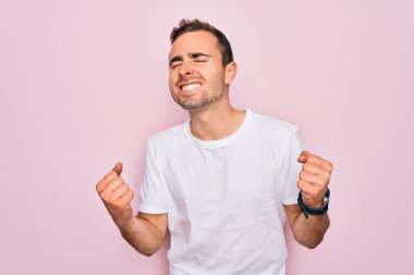 Mavi gözlü yakışıklı bir adam pembe arka planda sıradan beyaz tişörtler giyiyor çok mutlu ve heyecanlı bir şekilde kolları havada, gülümseyen ve başarı için çığlık atan bir jest yapıyor. Kutlama konsepti.