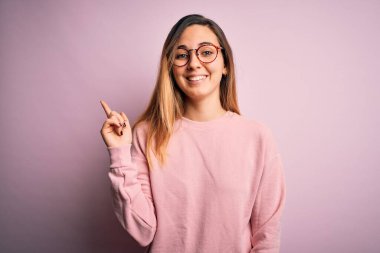 Güzel sarışın kadın, mavi gözlü, pembe arka planda gözlüklü, süveter giyen, yüzünde kocaman bir gülümseme olan, eli ve parmağı kameraya bakan..