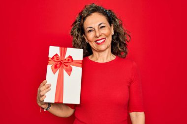 Orta yaşlı bir kadın elinde kırmızı izole edilmiş bir arka planda romantik bir hediye kutusu tutuyor mutlu bir yüzü var ve kendine güvenen bir gülümsemeyle dişlerini gösteriyor.