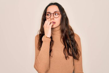Balıkçı yaka kazak giyen ve beyaz arka planda gözlük takan genç ve güzel bir kadın gergin görünüyor ve elleri ağızdan tırnak yiyor. Endişe sorunu.