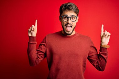 Sakallı, gözlüklü ve süveterli genç adam kırmızı arka planda dikilmiş şaşkın, şaşkın ve parmaklarıyla işaret ediyor ve kollarını kaldırıyordu..