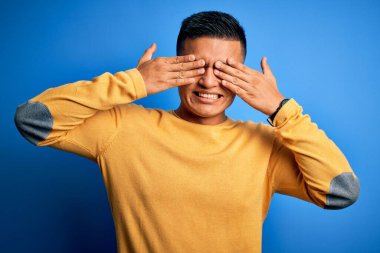 Sarı günlük süveter giyen genç, yakışıklı latin adam izole edilmiş mavi arka planda elleri gülücüklü ve komik bir şekilde gözlerini kapatıyor. Kör kavram.